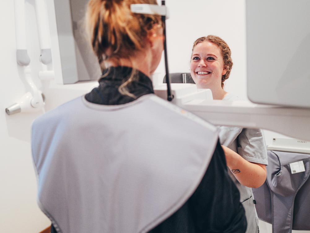 Eine Mitarbeiterin führt ein 3D-Röntgen bei einer Patientin durch. Die Mitarbeiterin lächelt die Patientin freundlich an und stellt dabei das Röntgengerät ein.