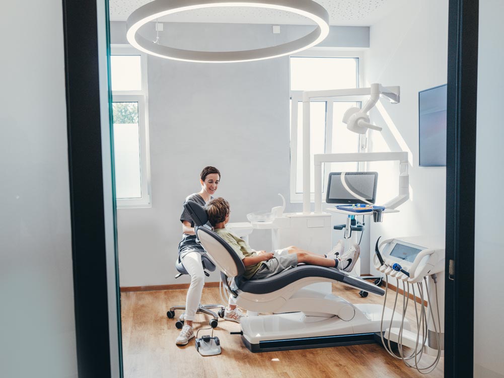 Zahnärztin Dr. Carolin Petsos unterhält sich lächelnd mit einem Kind im Behandlungsstuhl. Die Praxis ist hell und freundlich mit hellem Holzboden und moderner weiß-grauer Einrichtung.