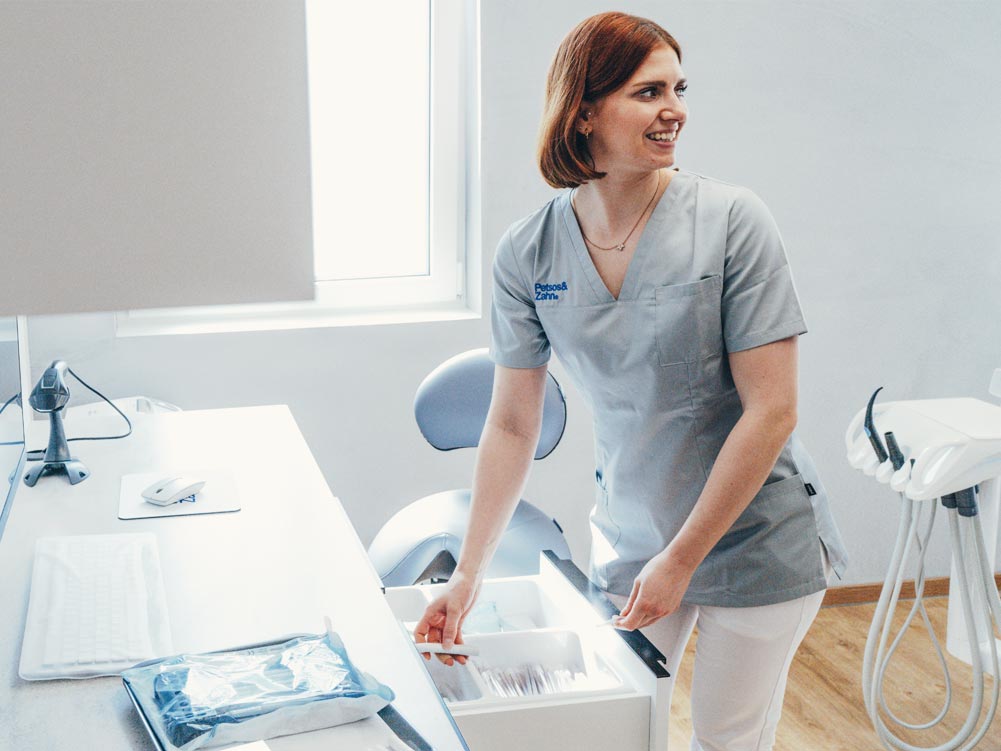 Eine Mitarbeiterin der Praxis Petsos&Zahn nimmt etwas aus einer Schublade im Behandlungszimmer. Sie schaut lachend eine Kollegin an, welche nicht im Bild zu sehen ist. Der Raum ist hell und freundlich mit moderner weiß-grauer Einrichtung und hellem Holzboden.