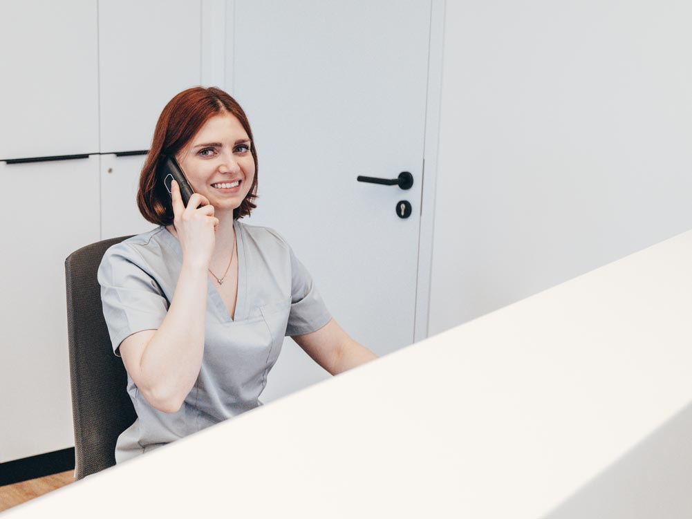 Eine herzliche Mitarbeiterin sitzt hinter dem Empfangstresen in der Praxis Petsos&Zahn und telefoniert mit einem Lächeln im Gesicht. Sie schaut freundlich in die Kamera.