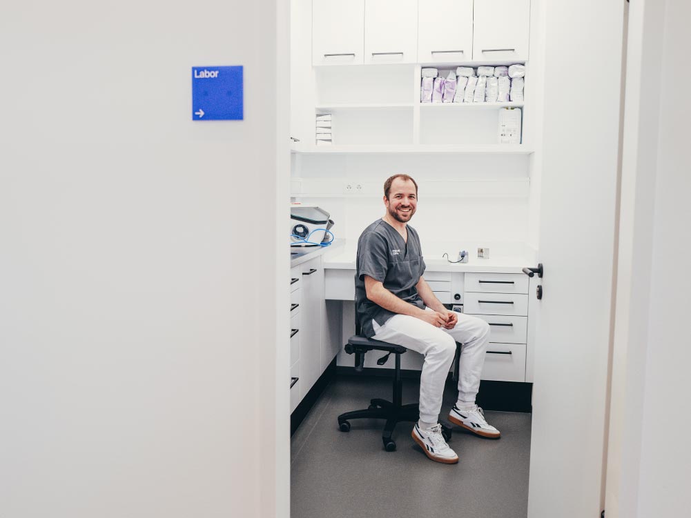 Zahnarzt Dr. Hari Petsos sitzt auf einem Stuhl in seinem praxiseigenen Dentallabor. Er lacht freundlich in die Kamera. Das Labor ist modern, hell und freundlich mit weißen Möbeln.