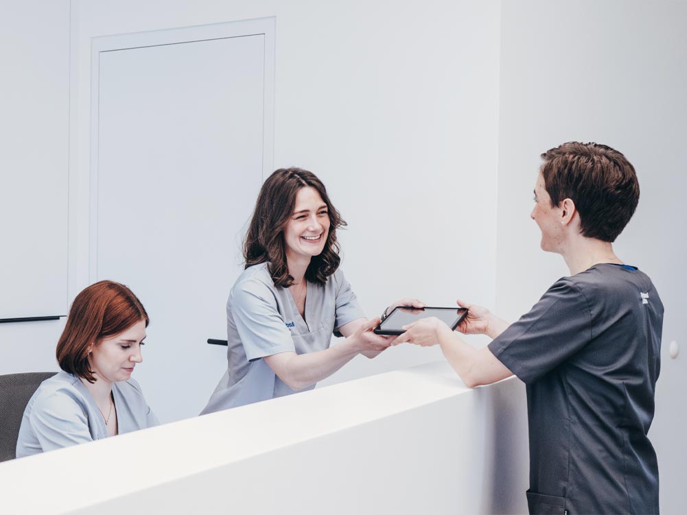 Eine herzlich lachende Mitarbeiterin am Empfang übergibt ein Tablet an Zahnärztin Dr. Carolin Petsos